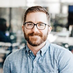 Professional headshot of Roy Cox wearing glasses and a blue shirt in an office environment